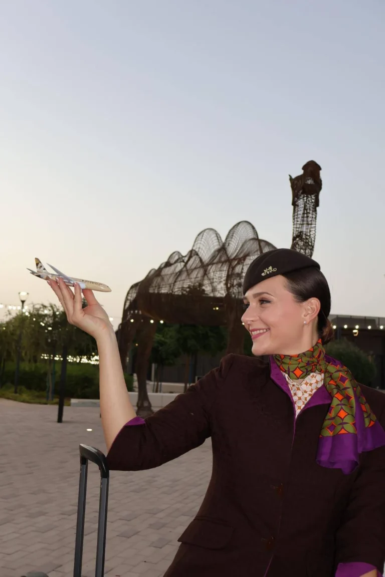 Letuška v uniforme Etihad Airways drží model lietadla pred veľkou drôtenou sochou ťavy v Abu Dhabi